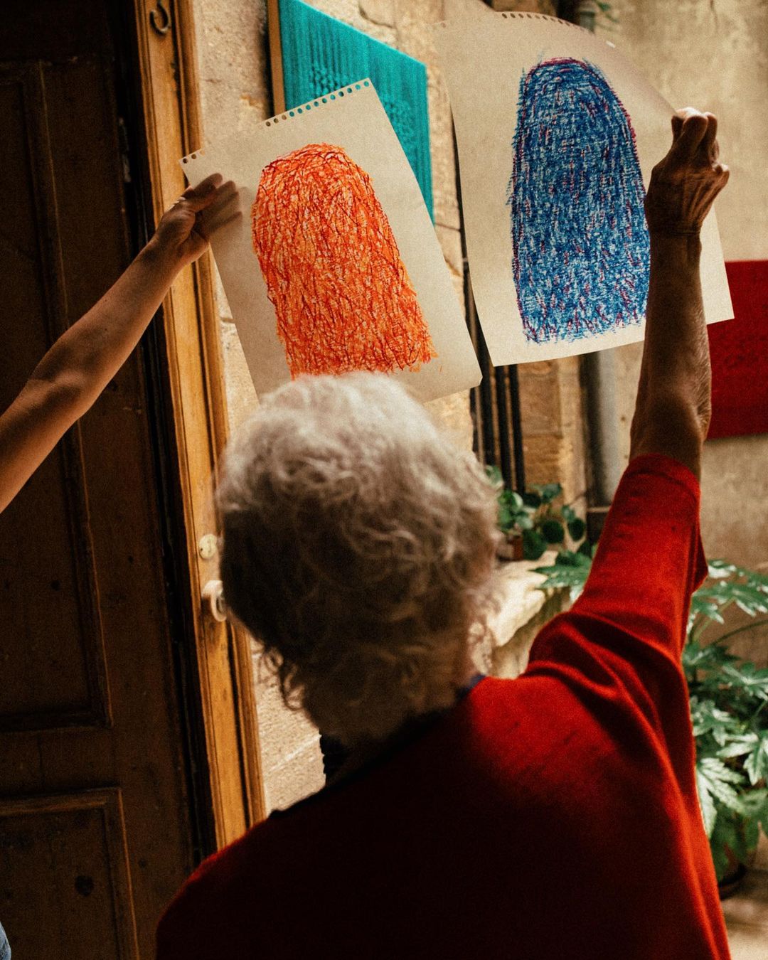 Two people holding crayon drawings of human silhouettes with scribbled texture on paper hung on a weathered building exterior.