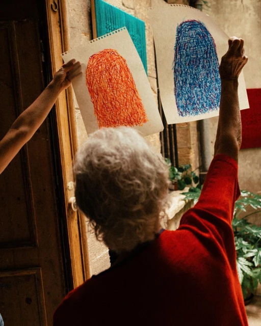 Two people holding crayon drawings of human silhouettes with scribbled texture on paper hung on a weathered building exterior.