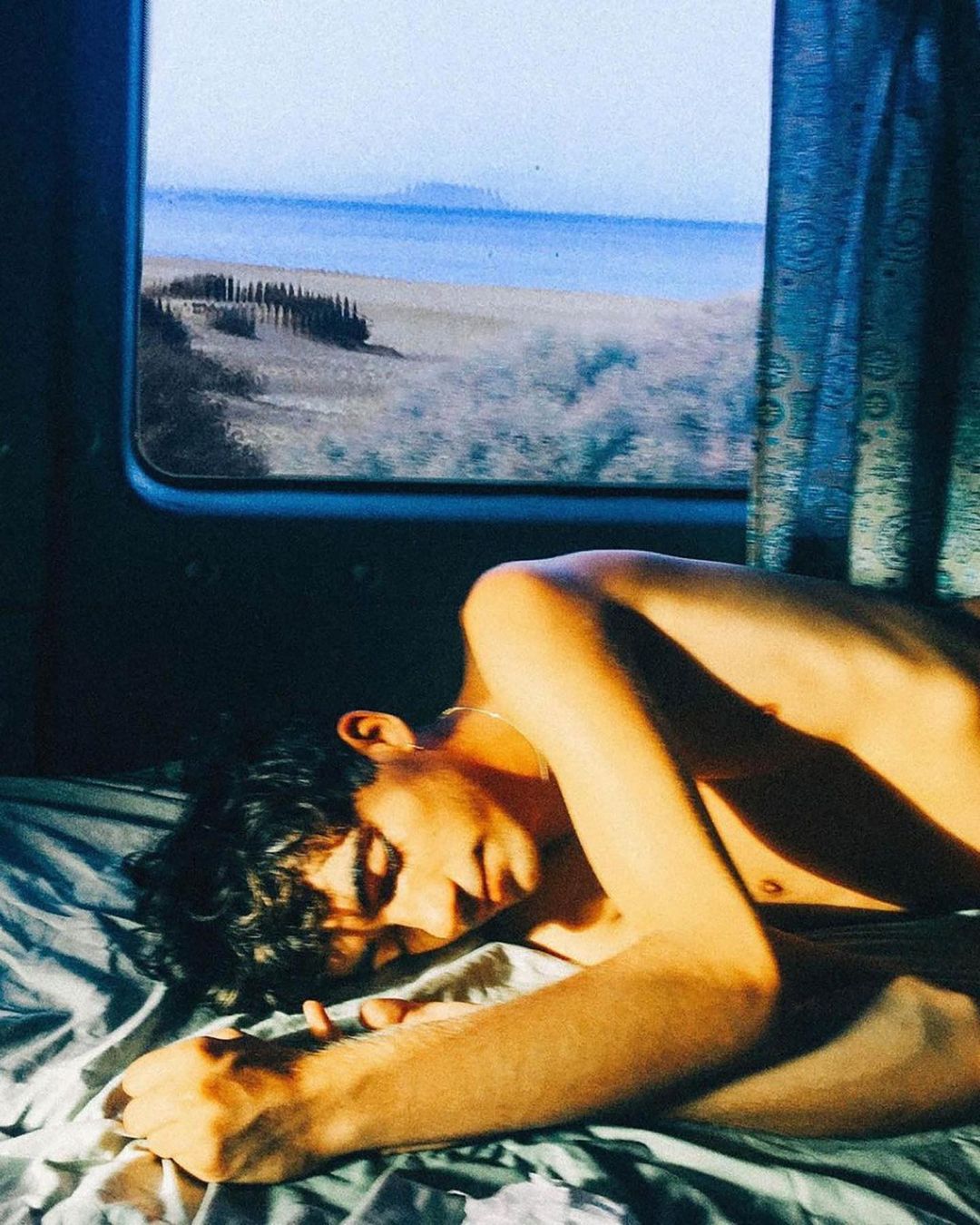 A young adult male with dark curly hair reclines on a bed in front of a window with a beach and ocean view in a close-up indoor shot.