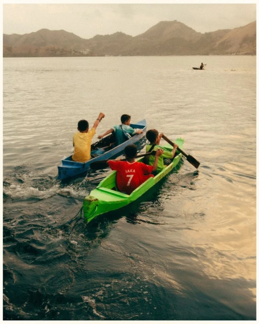Three young people rowing in brightly colored canoes on a lake, with mountains visible in the background under a cloudy sky at daytime.