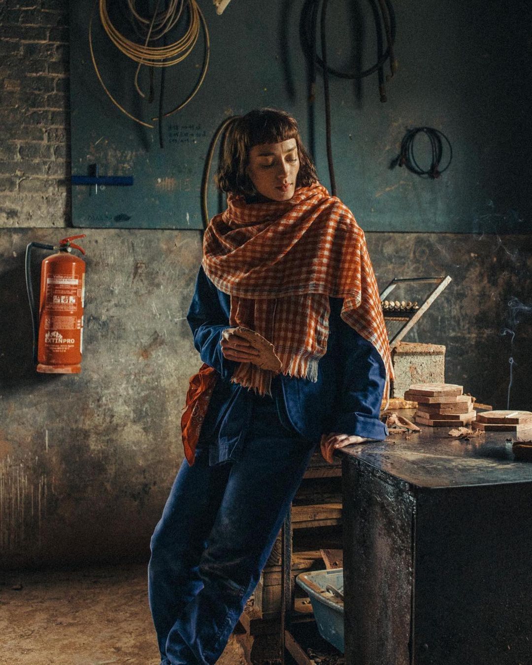 Woman in blue overalls and patterned scarf leans against a table in an industrial setting, with tools and equipment visible in the background.