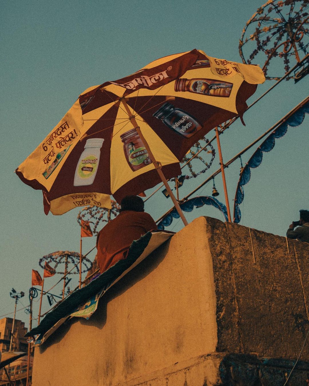 Person sits near a yellow and burgundy umbrella advertising a product; building and various decorative umbrellas in the background.