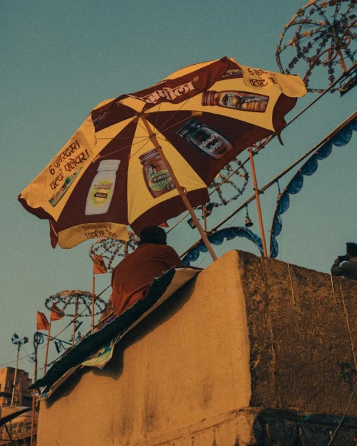 Person sits near a yellow and burgundy umbrella advertising a product; building and various decorative umbrellas in the background.