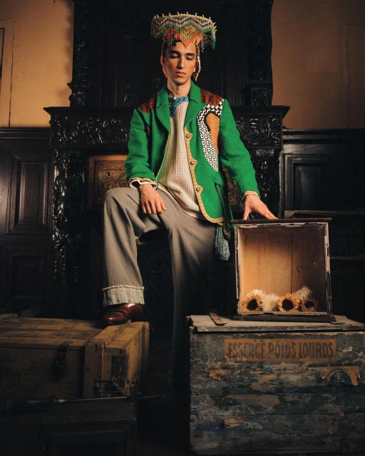 A male fashion model poses indoors wearing a green jacket, grey trousers, and a beaded headdress while standing by antique wooden boxes.