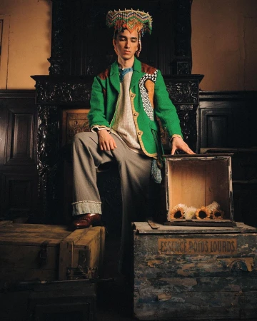 A male fashion model poses indoors wearing a green jacket, grey trousers, and a beaded headdress while standing by antique wooden boxes.