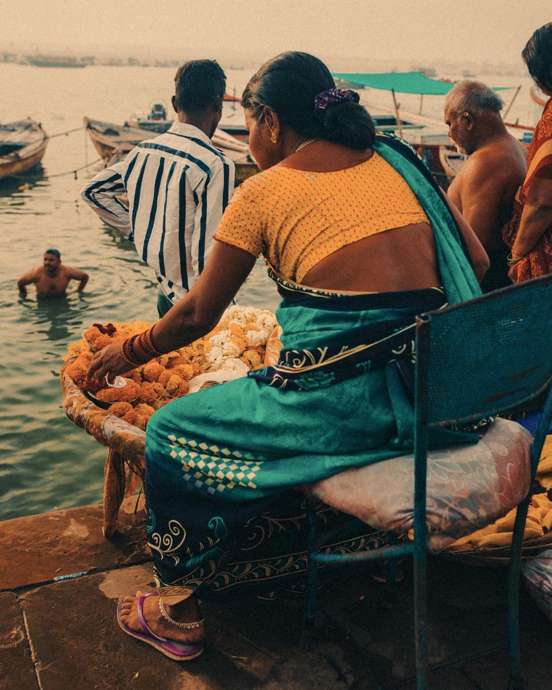 Woman in a yellow blouse and green sari sitting on a chair on a ghat next to flowers for sale, with boats on the river.