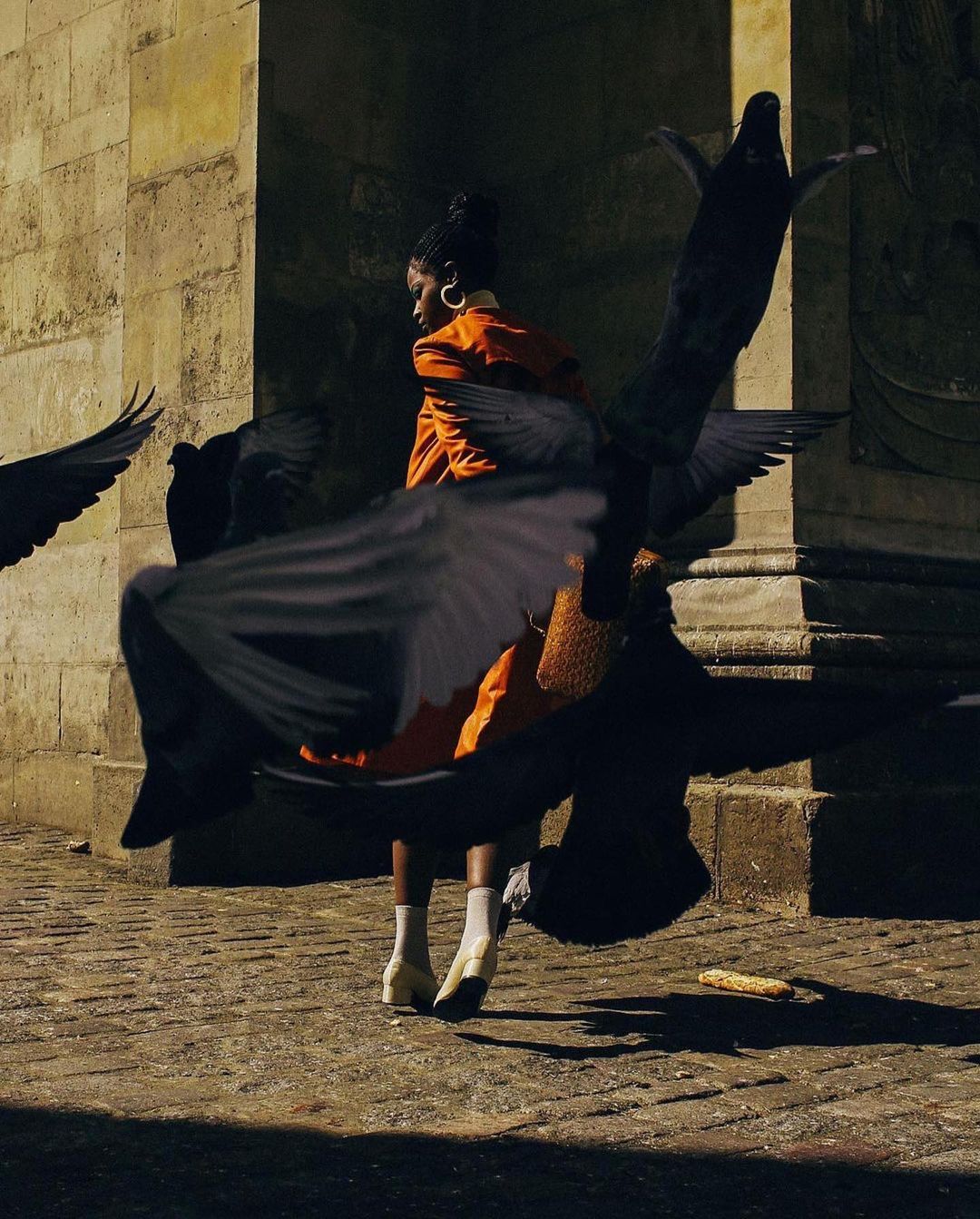 A stylish adult female in an orange suit surrounded by pigeons in an outdoor urban environment, enhanced by natural sunlight and a stone facade.