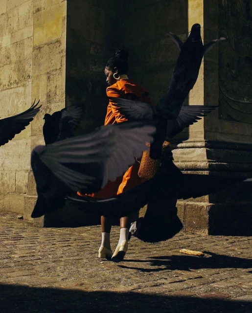 A stylish adult female in an orange suit surrounded by pigeons in an outdoor urban environment, enhanced by natural sunlight and a stone facade.