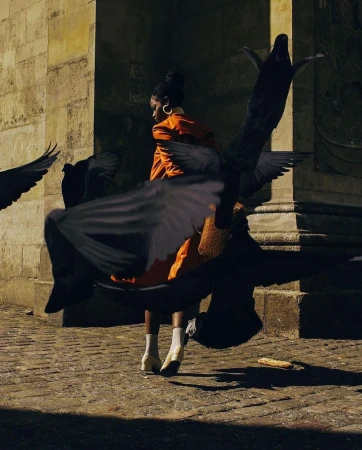 A stylish adult female in an orange suit surrounded by pigeons in an outdoor urban environment, enhanced by natural sunlight and a stone facade.
