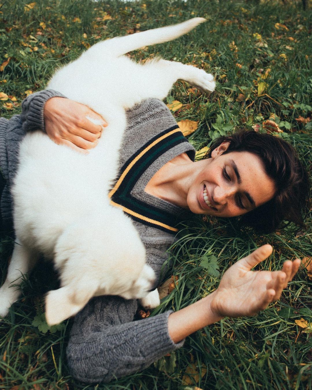 A smiling woman lies on her back in the grass, cuddling with a fluffy white puppy; they are both looking at the camera.
