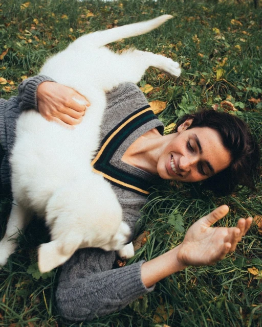 A smiling woman lies on her back in the grass, cuddling with a fluffy white puppy; they are both looking at the camera.