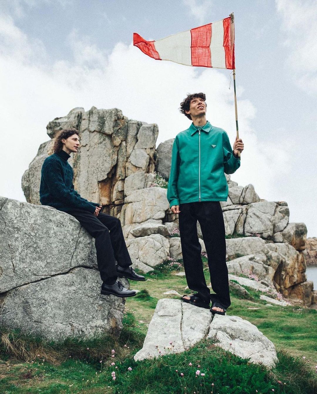 Two men pose with a flag on a rocky landscape with green grass and scattered clouds, creating a scenic outdoor fashion editorial.