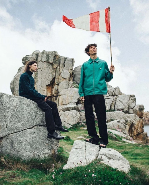 Two men pose with a flag on a rocky landscape with green grass and scattered clouds, creating a scenic outdoor fashion editorial.
