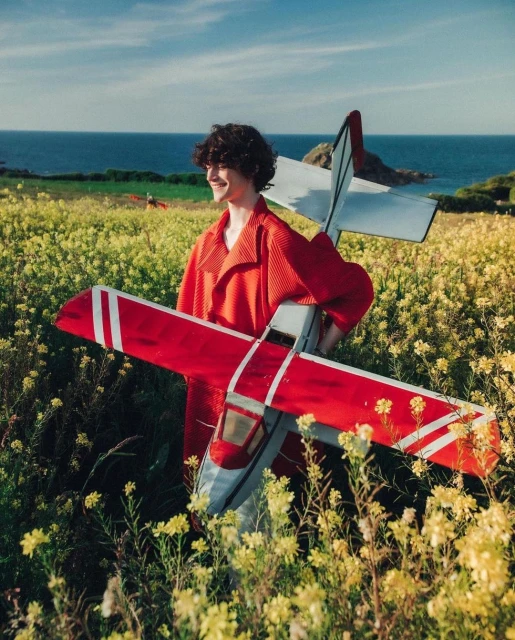 Teenager in orange jacket stands in field of yellow flowers holding red-and-white model airplane with ocean backdrop on sunny day.