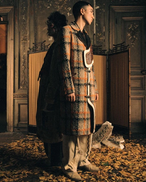 Two people pose indoors in a stylized fashion image. One is in focus wearing a patterned coat, while a second person stands behind.