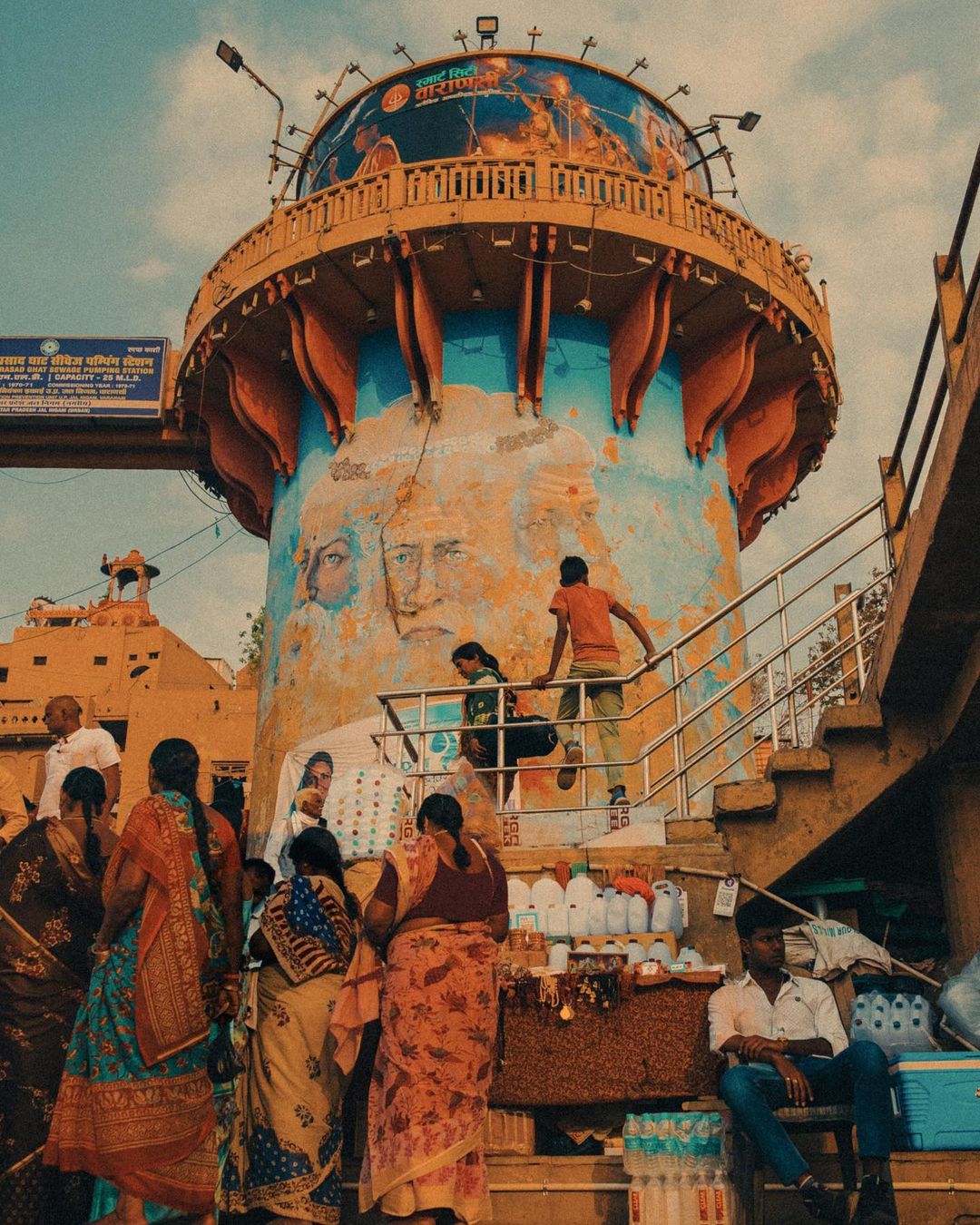 People gather around an ornate tower adorned with painted faces and murals, under a partly cloudy sky in an outdoor setting.
