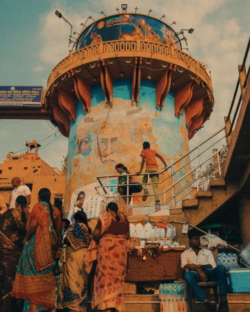 People gather around an ornate tower adorned with painted faces and murals, under a partly cloudy sky in an outdoor setting.