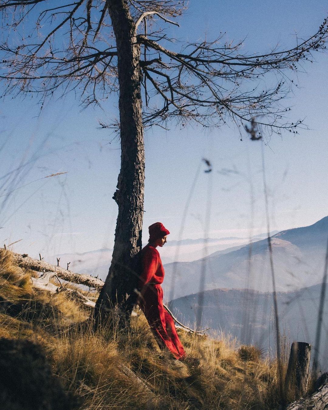 Adult woman in red leans against a bare tree on a hillside overlooking a mountain range with a hazy blue sky and tall grass.