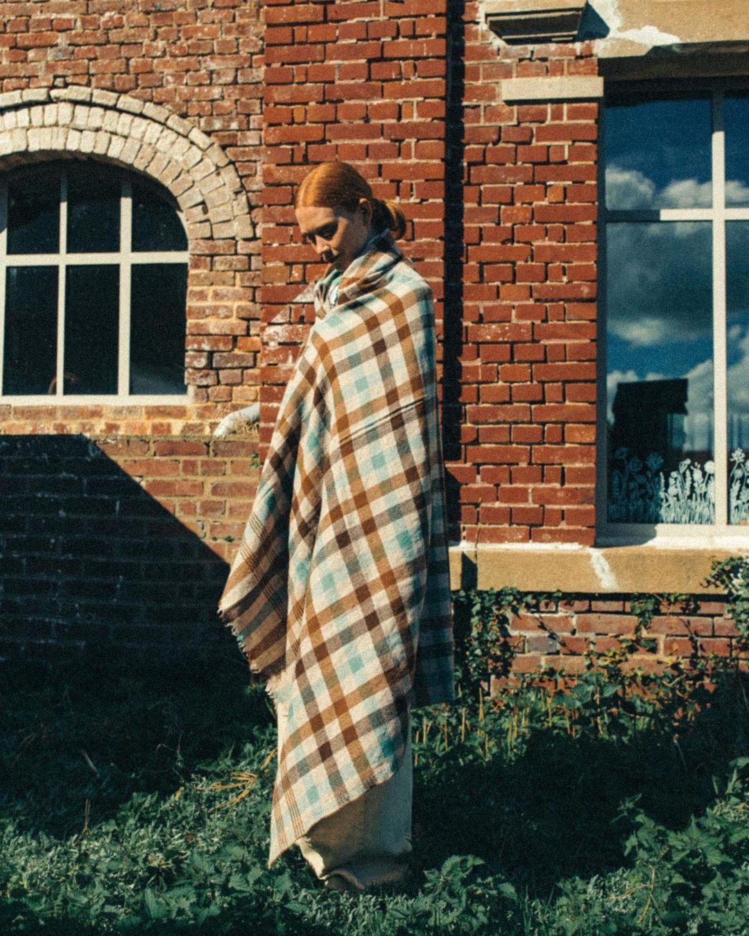 Red-haired woman draped in plaid blanket stands near brick building on a sunny day; plants in foreground; building reflection in window.