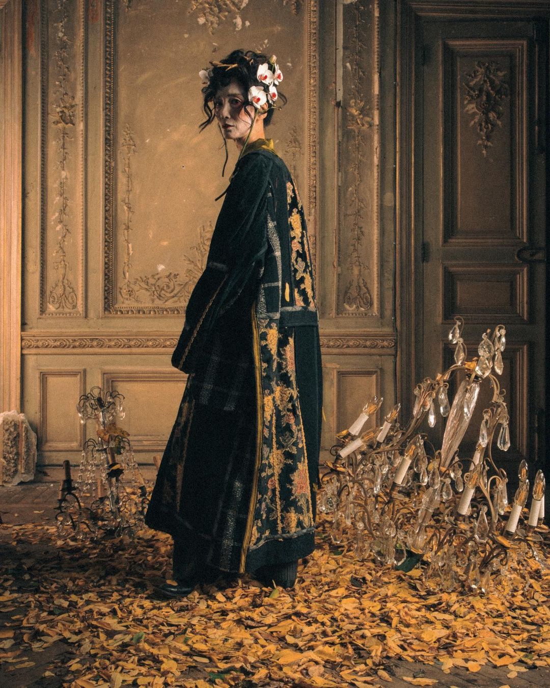 An adult female in a patterned coat stands inside a room with decorative moldings and gold leaves on the floor. A chandelier is visible.