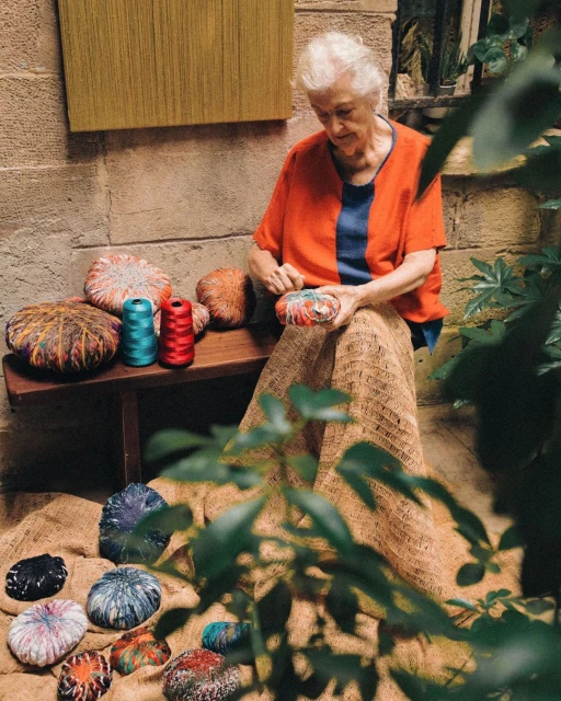 Elderly woman sits outside, surrounded by colorful yarn balls and spools. She is crafting with a needle in a tranquil outdoor setting.