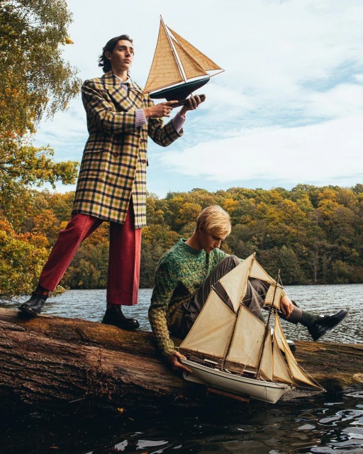 Two young men pose with model sailboats near a lake, featuring fall foliage, one standing in a plaid coat and the other sitting.
