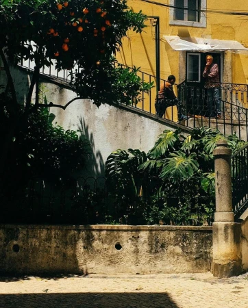 Two people standing on a balcony of a yellow building, with an orange tree in the foreground and cobblestone ground below.