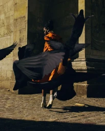 Black woman in orange coat and white socks walking past stone architecture amid a dramatic swarm of black pigeons in Paris.