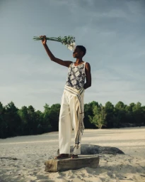 Black man standing on a stone slab on a sandy beach holding flowers up against an overcast sky.