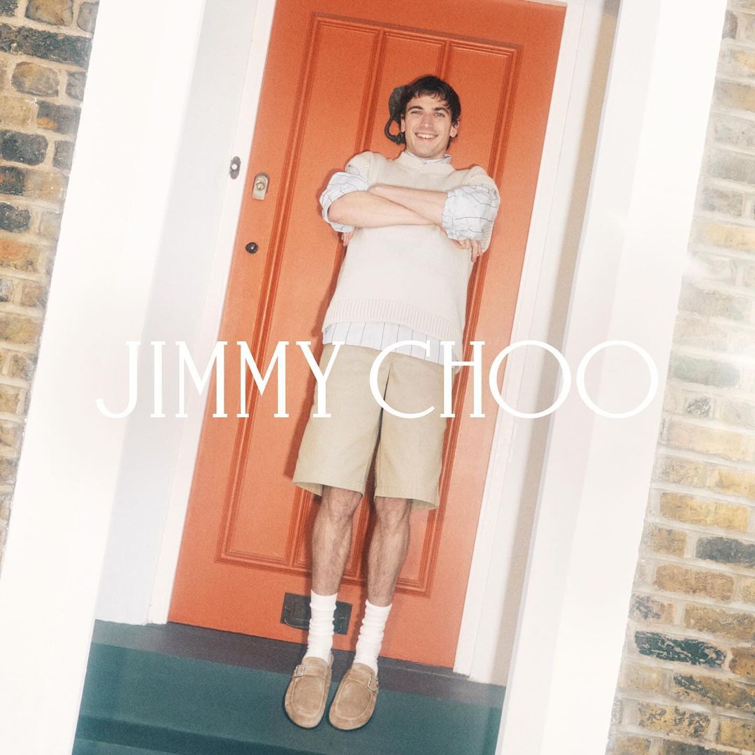 A young man in a casual outfit stands in a doorway with folded arms, posing for a Jimmy Choo fashion advertisement, outdoors in natural light.