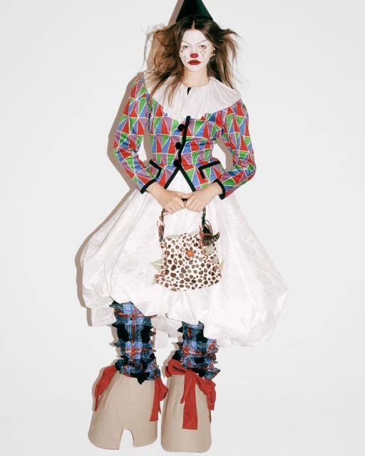 A fashion model wears a clown-inspired costume, including a colorful jacket, white dress, black hat, and oversized shoes, in a studio setting.
