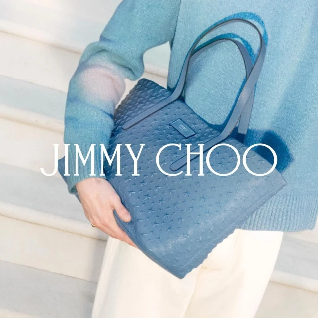 Fashion shot of a blue Jimmy Choo tote bag with star pattern held by a model wearing matching blue sweater and white pants.