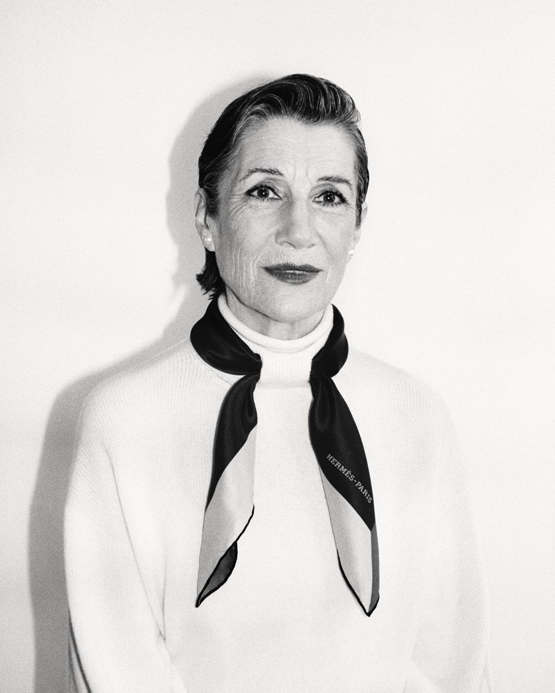 Elegant older woman in black-and-white portrait, wearing a cream turtleneck sweater and Hermès scarf, posing against a white background.