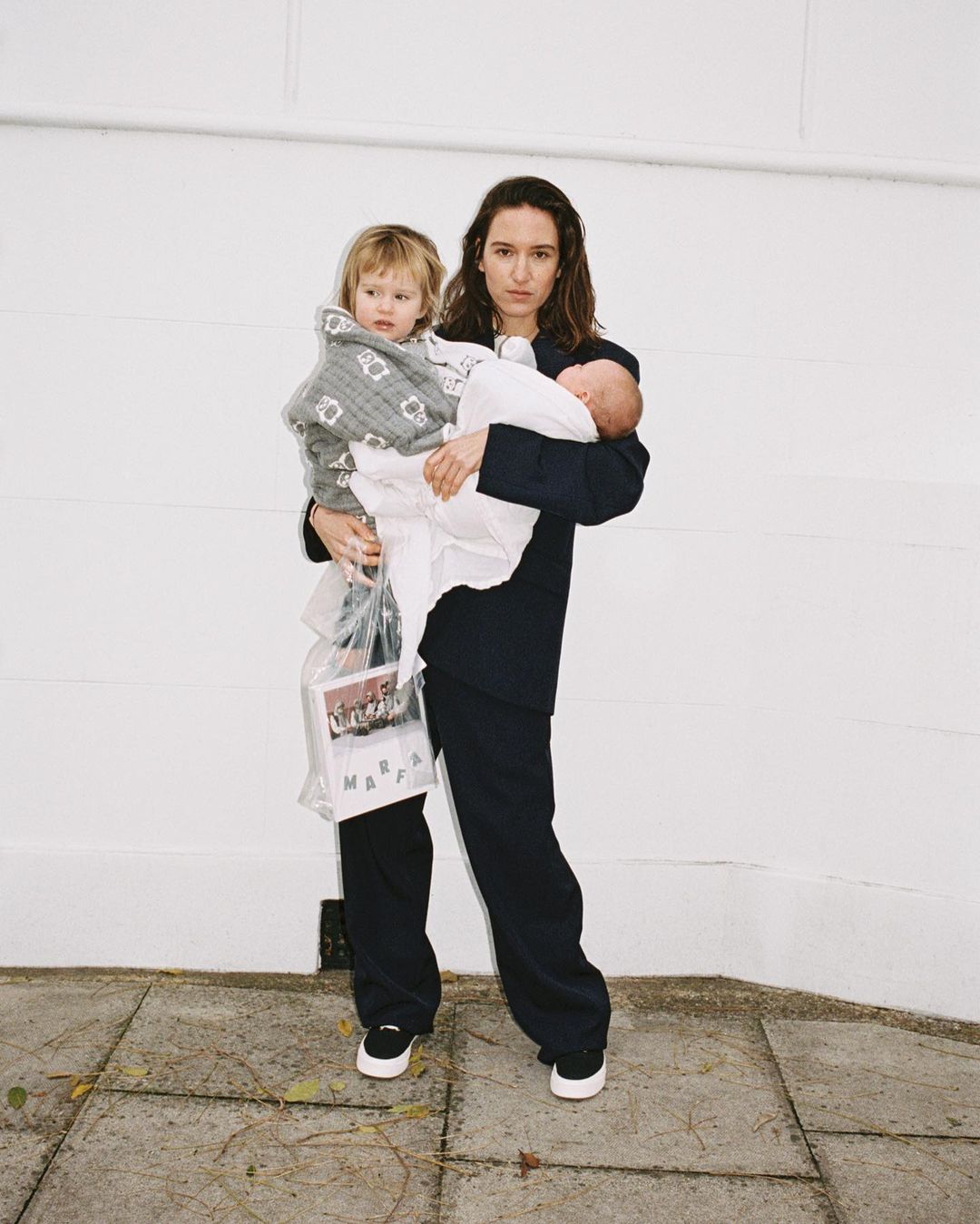 A mother in a dark suit stands outdoors holding a toddler wrapped in a patterned blanket and an infant wrapped in a white cloth.