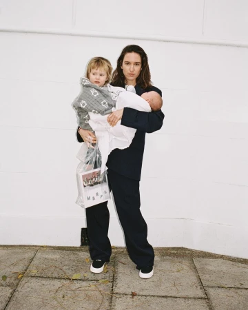A mother in a dark suit stands outdoors holding a toddler wrapped in a patterned blanket and an infant wrapped in a white cloth.