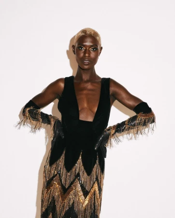 Model with blonde buzzed hair wearing a black dress with gold fringed gloves against a white background