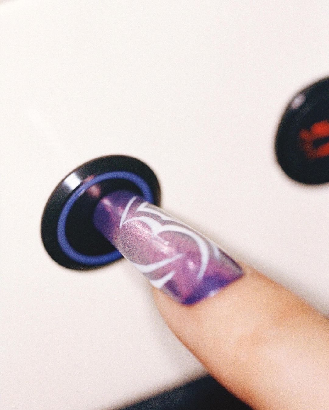 Close-up of a woman's hand with a long, square-shaped nail featuring a white swirl design over a purple ombre color scheme.