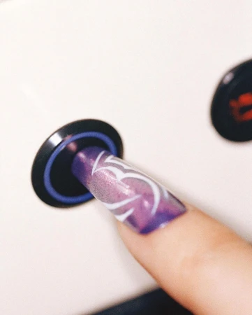 Close-up of a woman's hand with a long, square-shaped nail featuring a white swirl design over a purple ombre color scheme.