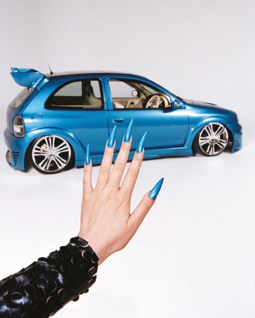 Custom blue hatchback car prominently displayed with a light skin hand and long, glossy blue nails, set against a clean white backdrop.
