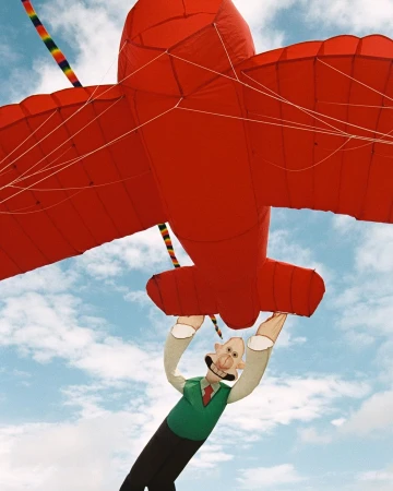 Red airplane kite with cartoon man attached is flown against a bright blue cloudy sky; outdoor activity and fun