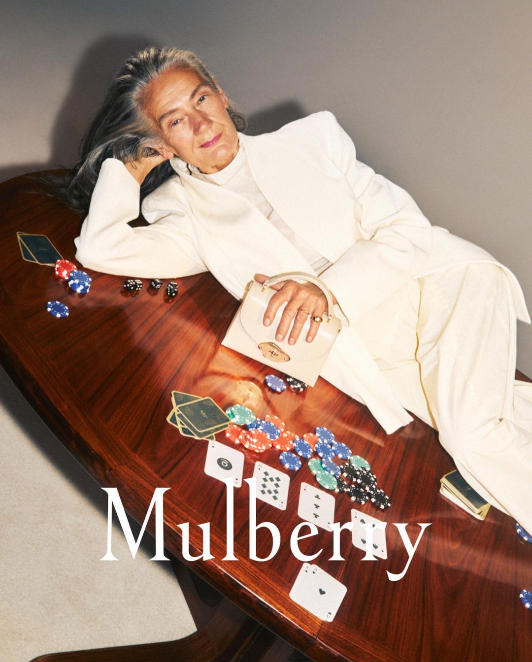 Mature woman in a white suit poses with a purse on a mahogany table, featuring scattered playing cards and poker chips in a fashion editorial.