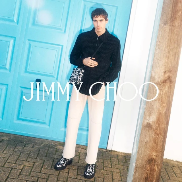 Front-facing shot of a male model wearing a black shirt, cream pants, and black and white shoes, with a Jimmy Choo bag.