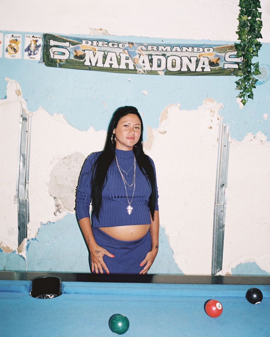 Adult female in a blue knit outfit posing in front of a light blue wall with a Diego Maradona banner and a blue pool table with billiard balls.