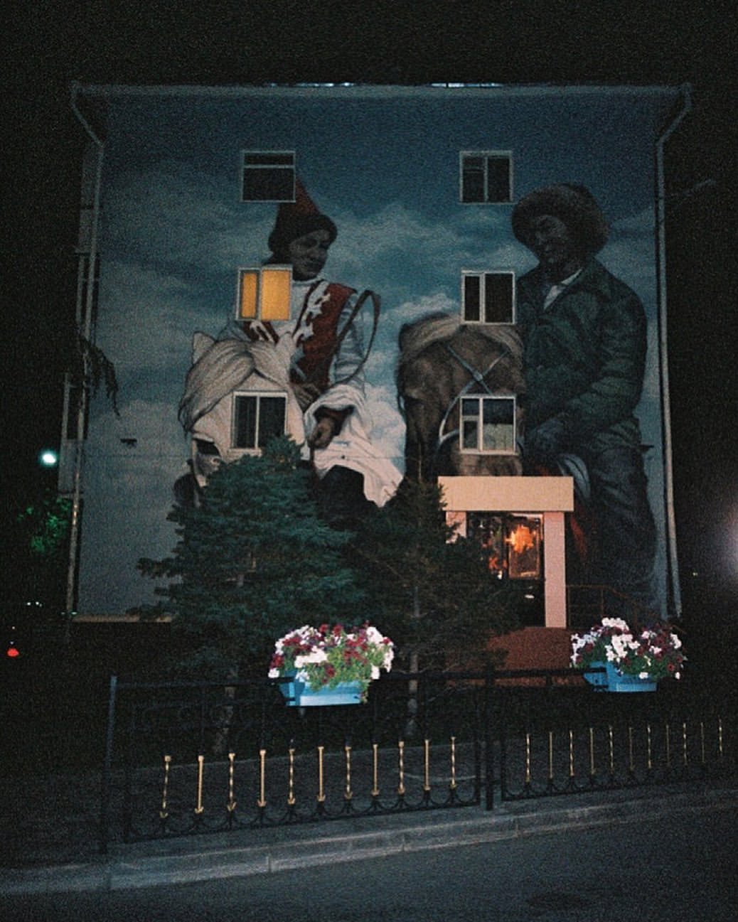Building exterior features a large mural with two figures on horseback, set against a cloudy sky, framed by trees and a fence with flower boxes.