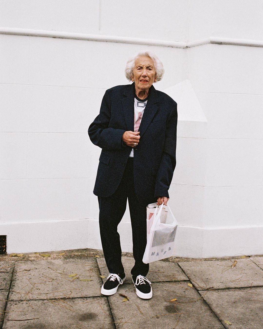 Elderly woman in oversized navy blazer and black sneakers stands against a white wall, holding a shopping bag on a tiled sidewalk.
