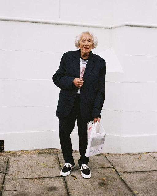 Elderly woman in oversized navy blazer and black sneakers stands against a white wall, holding a shopping bag on a tiled sidewalk.