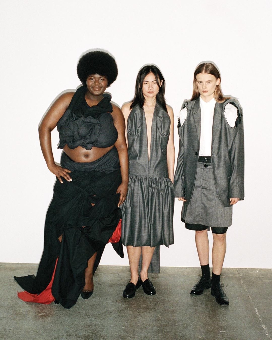 Three women pose in a studio wearing unconventional dark-colored garments with stylish designs, standing against a stark white backdrop.