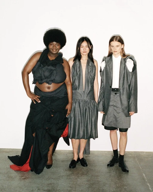 Three women pose in a studio wearing unconventional dark-colored garments with stylish designs, standing against a stark white backdrop.