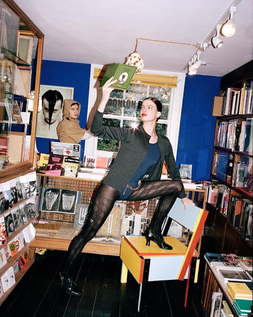 A fashionable woman strikes a pose on a chair in a bookstore while holding a book, with shelves and book covers lining the walls.