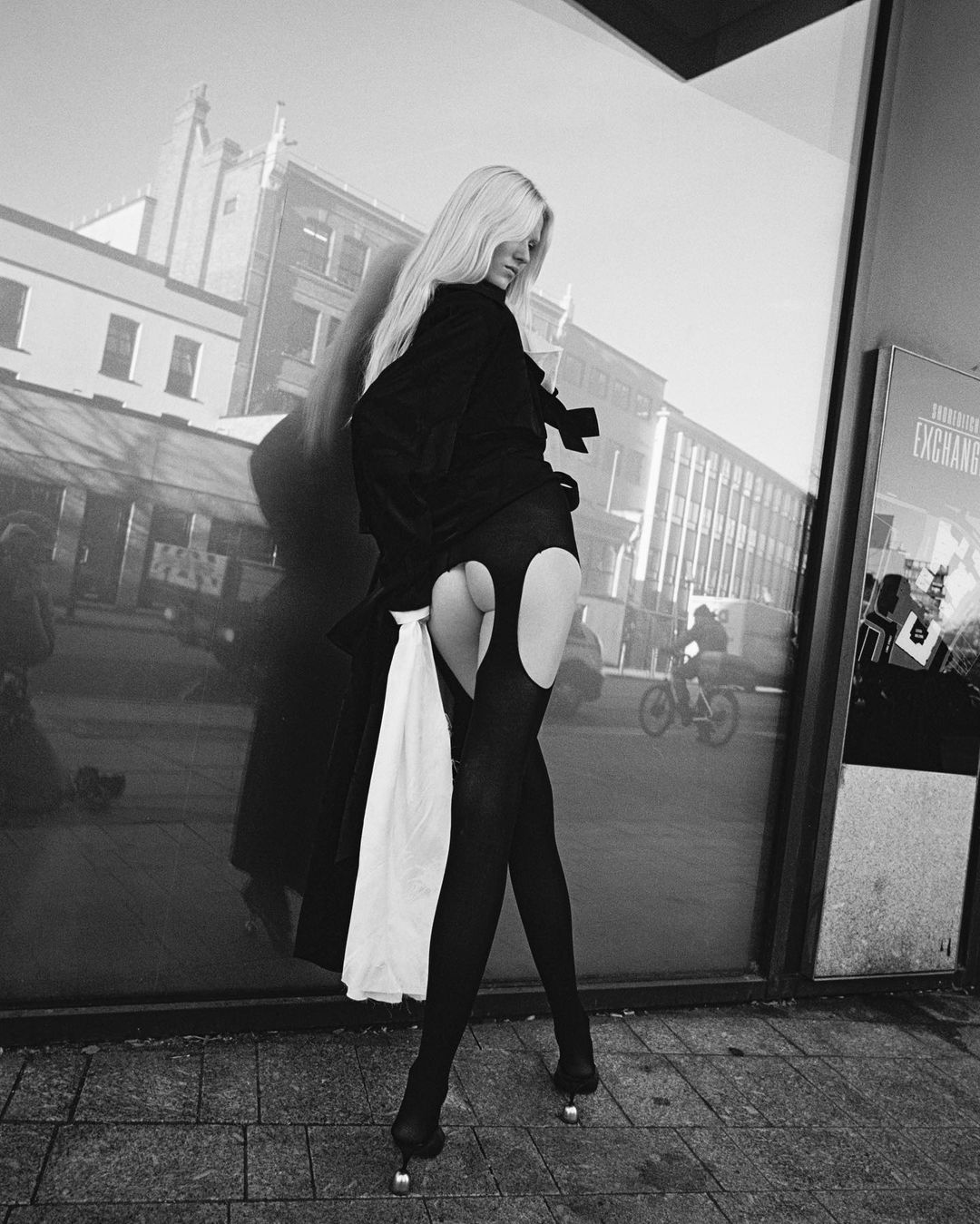 A person with long blonde hair poses in a city setting, wearing dark clothing and high heels, with a city scene reflected in glass.